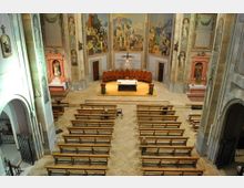 Interior de una iglesia con bancos de madera alineados hacia el altar elevado, donde hay una mesa central y un crucifijo. Las paredes del presbiterio están decoradas con grandes frescos religiosos y nichos laterales con estatuas.