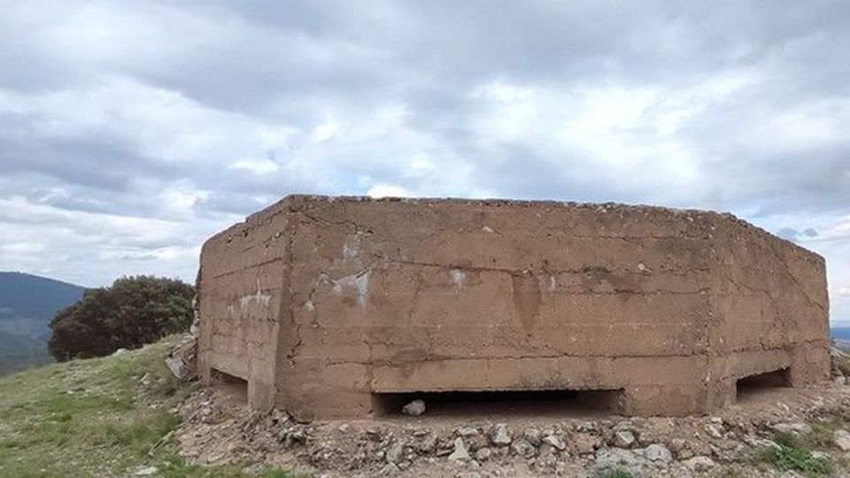 Búnker de hormigón rectangular y bajo, semienterrado en una loma cubierta de hierba y rocas, con una abertura horizontal en la base. Al fondo se ven colinas y un cielo nublado.