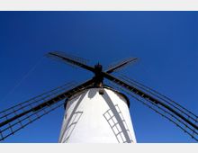 Vista en contrapicado de un molino de viento tradicional con torre cilíndrica blanca y grandes aspas en forma de cruz. El fondo es un cielo azul despejado, con sombras de las aspas proyectadas sobre la estructura.