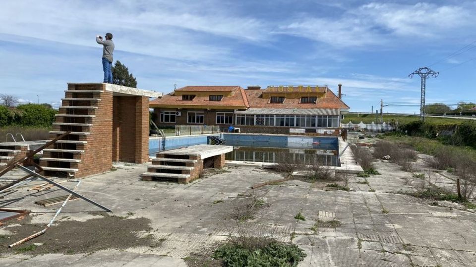 Zona exterior de un antiguo complejo con piscina vacía y deteriorada, rodeada de pavimento agrietado con maleza y restos de estructuras. En primer plano hay una plataforma y escaleras de ladrillo tipo trampolín, y al fondo un edificio alargado con tejado rojo y grandes ventanales bajo un cielo despejado.