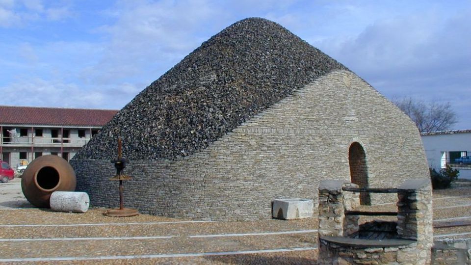 Edificio bajo de piedra con techo abovedado cubierto de grava oscura y una abertura arqueada, situado en una plaza pavimentada al aire libre. En primer plano hay un brocal de pozo de piedra y, al fondo, se ven otras construcciones y objetos de piedra bajo un cielo parcialmente nublado.