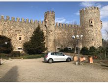 Muralla de piedra de un castillo o fortaleza con almenas y dos torres cilíndricas, vista desde una explanada de tierra. En primer plano hay un aparcamiento con un coche, farolas y jardineras, con árboles y cielo parcialmente nublado al fondo.