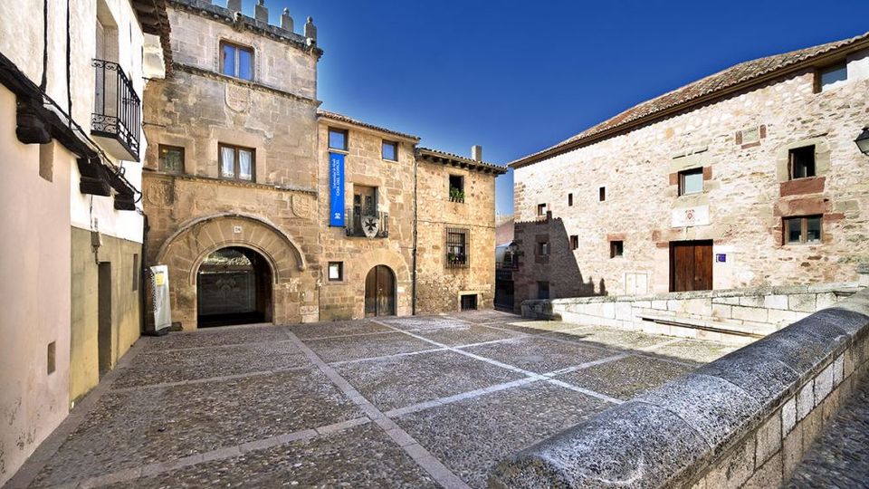 Plaza empedrada de un pueblo histórico, rodeada de edificios de piedra con arcos en la planta baja, ventanas pequeñas y tejados de teja. En primer plano se ve un murete de piedra y, al fondo, una torre con almenas bajo un cielo despejado.