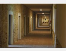 Pasillo interior largo con suelo de baldosas y paredes de ladrillo visto, con varias puertas alineadas a ambos lados y luces rectangulares en el techo. Al fondo se observa una zona con mobiliario y un extintor en la pared derecha.