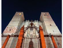 Fachada de una catedral de piedra vista desde abajo, con dos altas torres cuadradas iluminadas contra el cielo nocturno. En primer plano hay una reja de hierro forjado con un arco ornamentado y pilares de piedra anaranjada.
