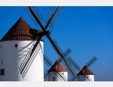 Molinos de viento tradicionales con torres cilíndricas blancas y techos cónicos marrones, con aspas de celosía negra superpuestas en primer plano. Al fondo se ven más molinos alineados bajo un cielo azul despejado.
