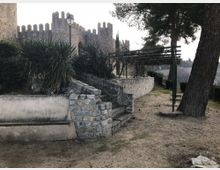 Jardín junto a una fortaleza de piedra con murallas almenadas, con un banco, escaleras y muros bajos de mampostería que rodean vegetación tipo agave. A la derecha hay una pérgola metálica sobre un sendero de tierra y varios árboles altos.