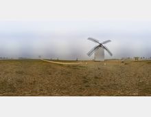 Panorámica de un terreno llano y pedregoso con un molino de viento tradicional blanco y aspas enrejadas en primer plano, bajo un cielo brumoso. Un sendero de tierra atraviesa el campo y, a lo lejos, se distingue otro molino y algunos elementos de señalización dispersos.