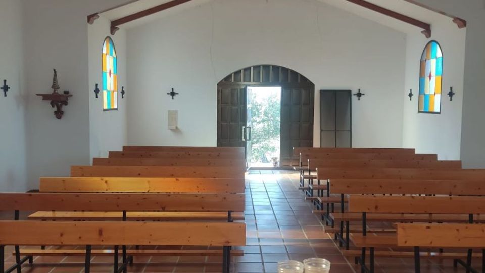 Interior de una pequeña capilla con bancos de madera alineados a ambos lados de un pasillo central sobre suelo de baldosas. Techo abovedado con vigas de madera vistas, vitrales laterales y una puerta doble al fondo que deja entrar luz natural.