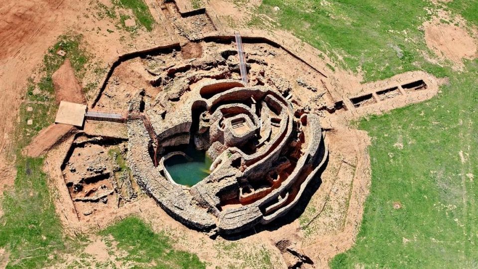 Vista aérea de un yacimiento arqueológico circular con muros concéntricos de piedra y una cavidad central con agua. Alrededor se ven zanjas de excavación y terreno mixto de tierra y pasto.