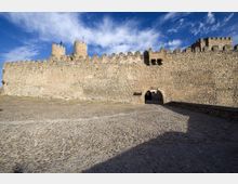 Muralla medieval de piedra con almenas y dos torres, con un arco de acceso en la parte inferior. Delante hay una amplia explanada empedrada y el fondo muestra cielo azul con nubes.