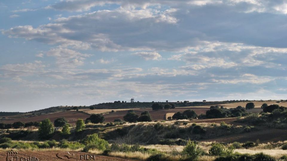 Paisaje rural de campos ondulados y matorral bajo, con parcelas de cultivo y árboles dispersos. En el horizonte se distingue un molino de viento sobre una colina bajo un cielo amplio con nubes.