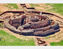 Vista aérea de un yacimiento arqueológico con muros de piedra concéntricos que forman recintos circulares y pasillos, parcialmente excavados. Alrededor se observa terreno de tierra y pasto con zanjas de excavación, estacas perimetrales y pasarelas metálicas de acceso.