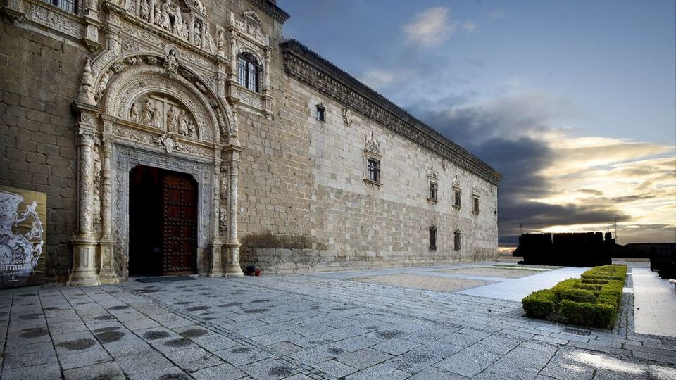 Fachada de un edificio histórico de piedra con una portada monumental esculpida y arco decorado junto a una gran puerta de madera. Delante hay una amplia explanada pavimentada y setos recortados, con el cielo nublado al fondo.