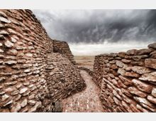 Pasaje estrecho entre muros de piedra apilada, con suelo de tierra, que conduce hacia un paisaje desértico abierto. Al fondo se ve un horizonte plano bajo un cielo nublado y oscuro.