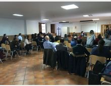 Sala de reuniones con suelo de baldosas y paredes blancas con panelado de madera, iluminada por plafones rectangulares en el techo. Filas de sillas orientadas hacia una pizarra y una puerta al fondo, con varias ventanas laterales.
