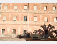 Fachada de un edificio antiguo de varias plantas con ladrillo visto y zonas de piedra, con ventanas rectangulares y algunas rejas en la planta baja. En primer plano hay un pequeño patio pavimentado y se proyecta la sombra de una palmera sobre la pared.