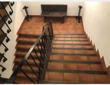Escalera interior con peldaños de baldosas de terracota y barandillas de hierro negro, que desciende a un rellano. En el rellano hay un banco de madera contra la pared blanca y dos faroles decorativos a los lados.