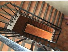 Vista cenital de una escalera interior en espiral cuadrada, con peldaños de baldosas de terracota y barandillas de hierro negro. El hueco central muestra varios tramos descendentes enmarcados por paredes claras y pasamanos metálicos.