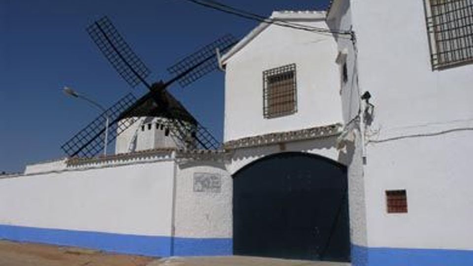 Casa encalada con zócalo azul y gran portón arqueado negro, con ventanas enrejadas y tejado de tejas. Detrás se eleva un molino de viento con aspas de celosía bajo un cielo despejado.