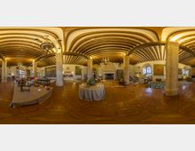 Gran salón interior de estilo histórico con suelo de piedra, columnas de piedra y techo abovedado con vigas de madera y lámparas de araña. Al fondo hay una gran chimenea, tapices en las paredes y varias mesas y zonas de estar distribuidas por la sala.