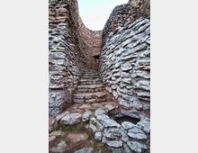Pasaje estrecho de piedra con escalones irregulares, flanqueado por altos muros de mampostería rugosa que se curvan hacia una abertura al cielo. En primer plano se aprecia un pequeño hueco circular en el suelo de piedra.