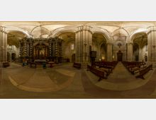 Interior de una catedral de piedra con altas bóvedas de crucería y grandes columnas, con filas de bancos de madera en la nave. A un lado se ve un retablo barroco oscuro con columnas salomónicas y una reja, junto a un púlpito y vidrieras.