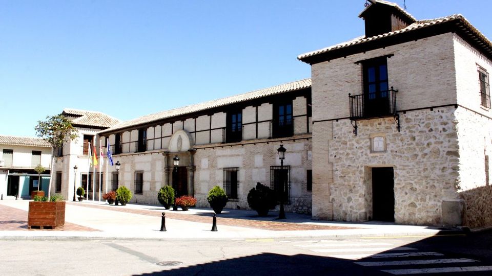 Edificio institucional de dos plantas con fachada de piedra y ladrillo, detalles de entramado oscuro y una torre lateral con pequeño balcón. Frente a la entrada hay una plaza pavimentada con farolas, maceteros y varias banderas, junto a un paso de cebra en la calle.