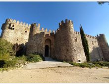 Castillo de piedra con murallas almenadas y torres cilíndricas, con una puerta de arco en el centro accesible por una rampa. En primer plano hay un camino de tierra y vegetación baja, con un ciprés junto al muro bajo un cielo despejado.