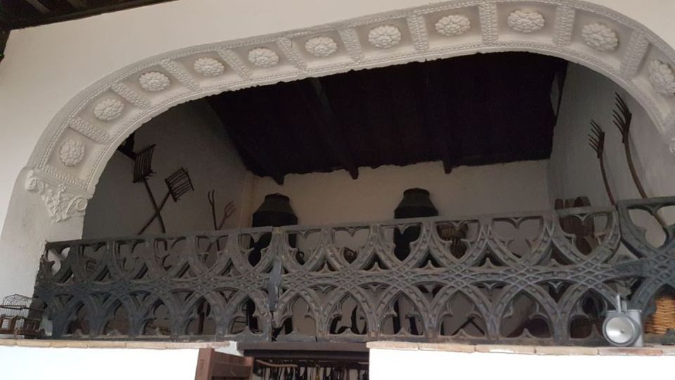Interior de un edificio tradicional con un gran arco de yesería decorado con relieves y una barandilla de madera tallada en un altillo. Al fondo se ve un techo de vigas oscuras y paredes con herramientas agrícolas colgadas.