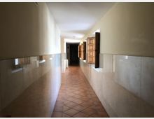 Pasillo interior largo y estrecho con suelo de baldosas de terracota en diagonal y paredes claras con revestimiento cerámico hasta media altura. A la derecha hay ventanas pequeñas con contraventanas de madera abiertas que dejan entrar luz natural hacia el fondo del corredor.