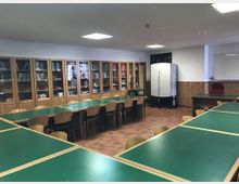 Sala de estudio o aula con mesas largas de superficie verde y sillas de madera, sobre suelo de baldosas. Al fondo hay estanterías acristaladas con libros, paredes con revestimiento de madera, iluminación de techo y una pizarra en un lateral.