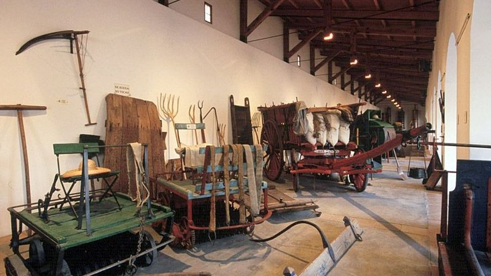 Interior de un museo o almacén histórico con techo alto de vigas de madera y una larga pared blanca. A lo largo del pasillo se exhiben carros y herramientas agrícolas antiguas, incluidos arados, trineos y carretas de madera y metal sobre el suelo de cemento.