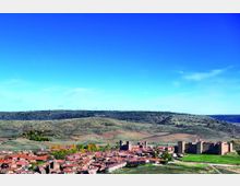 Vista panorámica de un pueblo con tejados rojos en un valle, junto a un gran castillo amurallado con torres y una iglesia destacada. Al fondo se ven colinas y campos cultivados bajo un cielo azul despejado.