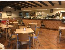 Interior de una cafetería-restaurante con suelo de baldosas de barro, mesas cuadradas y sillas de madera y metal. Al fondo hay una barra larga revestida de azulejos con vitrina y estanterías, y el techo muestra vigas oscuras vistas e iluminación empotrada.