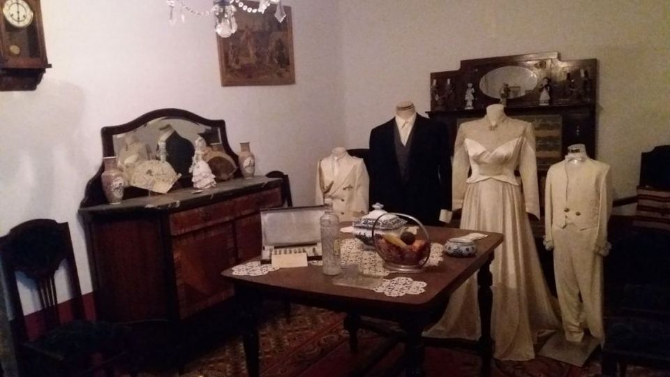 Sala de museo con muebles antiguos de madera, mesa central con tapetes de encaje y objetos decorativos, y aparadores con espejos. Varias figuras de exhibición muestran trajes formales de época, bajo una lámpara de araña y junto a un reloj de pared.
