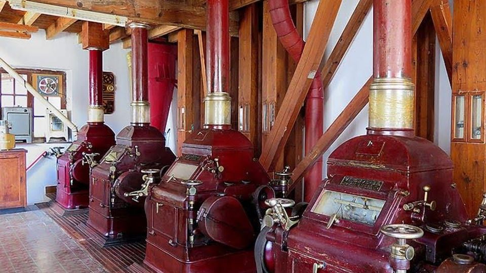Sala industrial con grandes máquinas antiguas de metal color rojo alineadas, cada una con una alta chimenea cilíndrica y tuberías, bajo un techo de vigas de madera vista. El espacio tiene suelo de baldosas y estructura interior de madera con refuerzos diagonales.