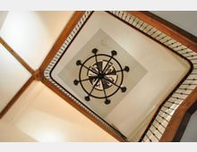 Vista hacia arriba de un hueco de escalera con barandillas de metal y vigas de madera, formando un marco cuadrado alrededor del techo. En el centro hay una lámpara colgante circular de hierro sobre un panel decorativo.