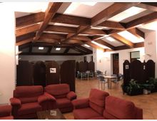 Salón interior tipo cafetería con techo alto de vigas de madera vistas y lucernarios, paredes blancas y suelo de baldosas. En primer plano hay sofás rojos y, al fondo, mesas con sillas separadas por biombos oscuros y algunas plantas junto a una puerta de madera.