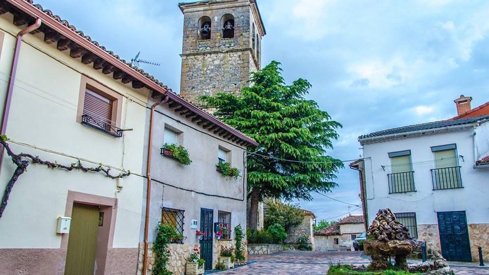 Plaza de un pueblo con pavimento de adoquines, rodeada de casas bajas de fachadas claras y tejados de teja roja. Al fondo destaca una torre de iglesia de piedra con campanario y cruz, junto a un árbol grande y una pequeña fuente o escultura de roca en un parterre circular.