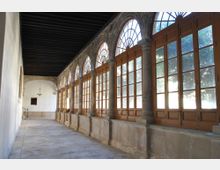 Galería interior tipo claustro con suelo de piedra y techo de vigas de madera oscura, a lo largo de una pared blanca. En el lateral derecho hay una serie de arcos de piedra con grandes ventanales y puertas de madera acristaladas que dejan entrar luz y muestran árboles en el exterior.