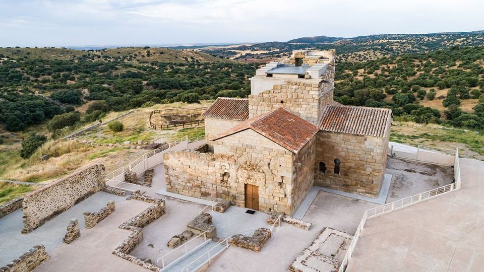 Iglesia de piedra en una colina, con tejados de teja y una torre cuadrada, rodeada por un recinto vallado y ruinas arqueológicas de muros bajos. Al fondo se extiende un paisaje de dehesa con colinas y árboles dispersos bajo cielo abierto.