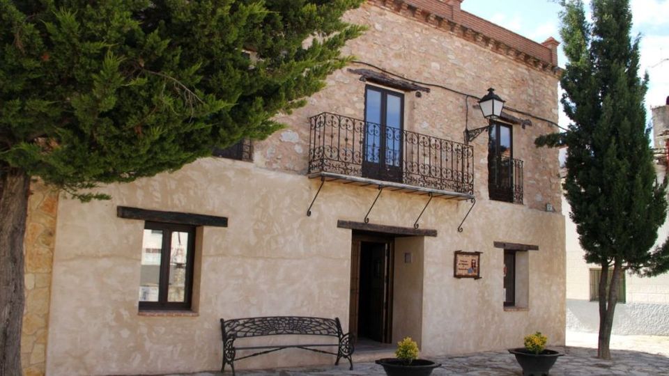 Fachada de una casa rural de dos plantas con muros de piedra y revoco claro, balcón de hierro forjado y puerta central bajo una farola. Delante hay una pequeña plaza empedrada con un banco metálico, maceteros y árboles a ambos lados.