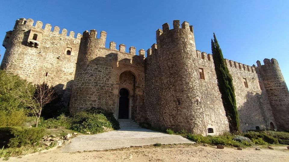 Castillo de piedra con murallas almenadas y torres cilíndricas, con una puerta de arco en el centro accesible por una rampa. En primer plano hay un camino de tierra y vegetación baja, con un ciprés junto al muro bajo un cielo despejado.