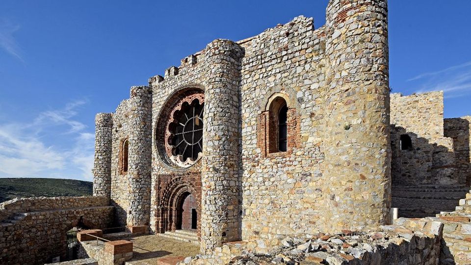 Fachada de una iglesia o monasterio medieval de piedra en ruinas, con contrafuertes cilíndricos, portal arqueado y un gran rosetón circular. El edificio se alza sobre una zona elevada con muros de piedra y un sendero, bajo un cielo azul despejado.