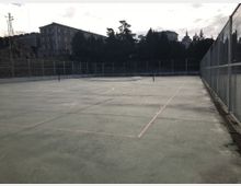 Pista deportiva exterior de hormigón con líneas marcadas y postes para red en el centro, rodeada por vallas metálicas altas. Al fondo se ven árboles y edificios en una zona elevada bajo un cielo nublado.