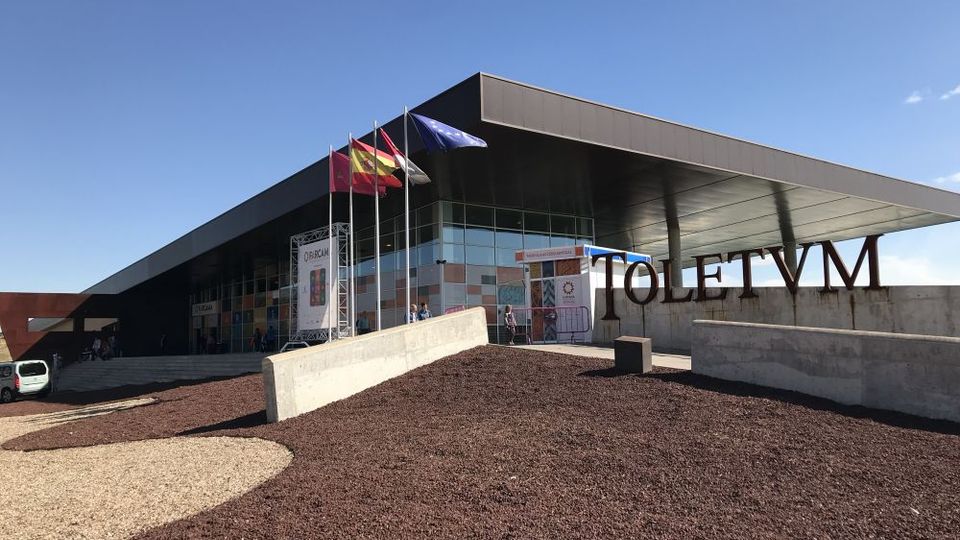 Entrada de un edificio moderno con gran marquesina metálica y fachada acristalada, frente a una explanada ajardinada con grava y muros de hormigón. En primer plano aparecen letras grandes que forman “TOLETVM” y varios mástiles con banderas junto al acceso.