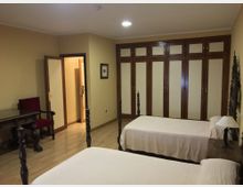 Dormitorio con dos camas individuales de estructura de madera oscura y colchas blancas, suelo de madera clara y paredes beige. Al fondo hay un gran armario empotrado con puertas de madera y paneles claros, y a un lado un escritorio con silla junto a una puerta abierta hacia el pasillo.